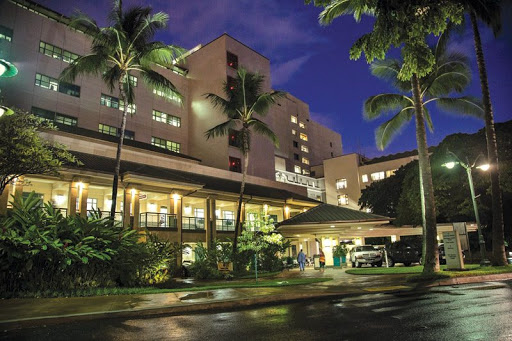 Exterior of The Queen's Medical Center at night with palm trees and other tropical landscaping.