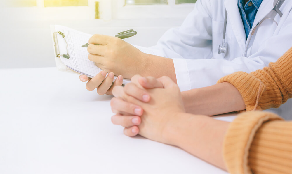 Hands of a doctor filling out paperwork next to clasped hands of a patient.