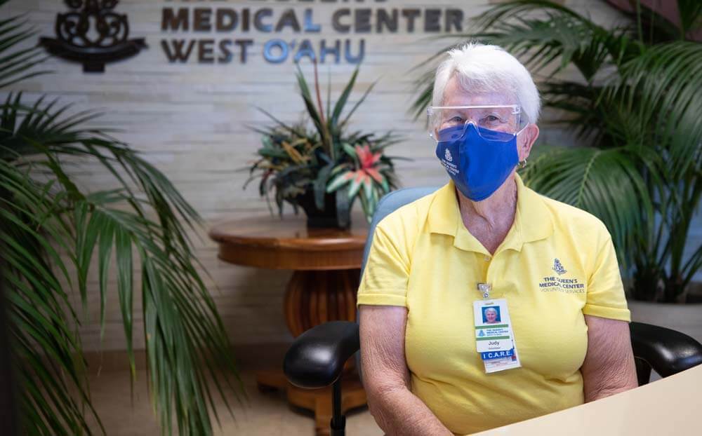 Woman in yellow polo and blue face mask sitting at reception desk.
