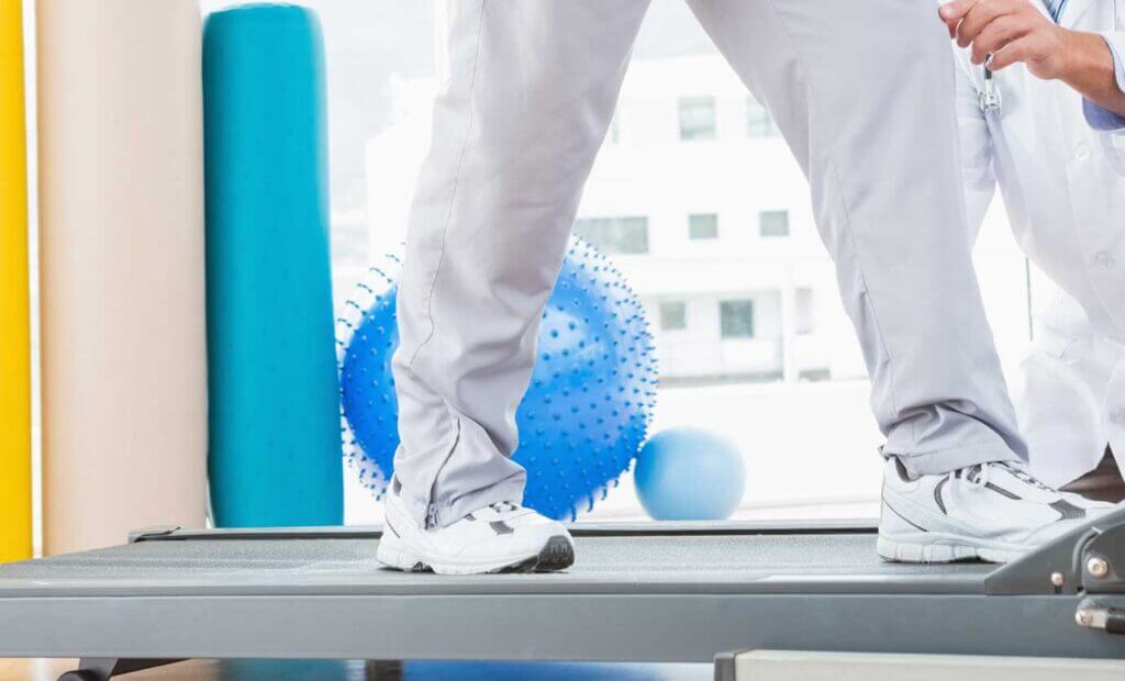 Close up of legs walking on a treadmill with physician therapy supplies in the background.