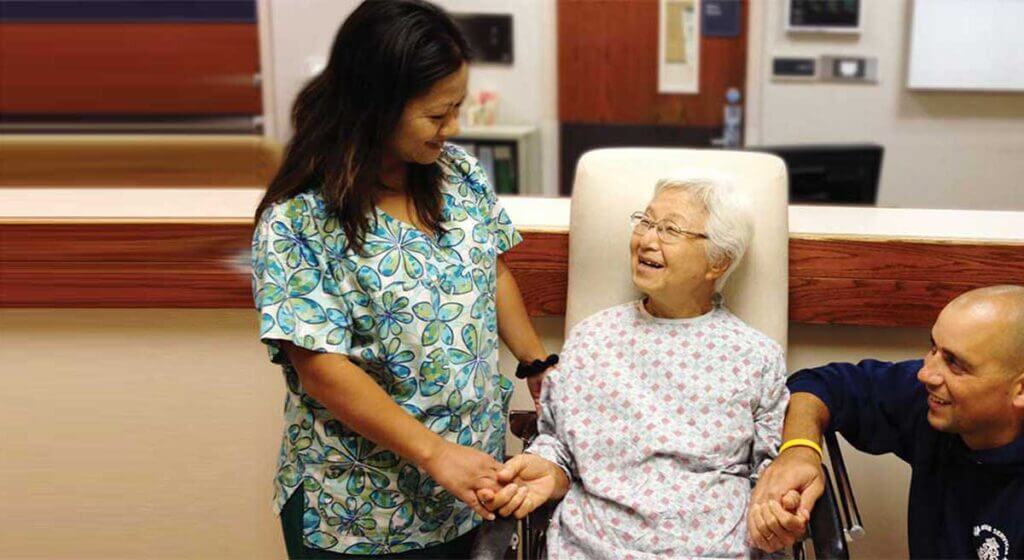 2 nurses laugh and hold the hands of an elderly patient in chair.